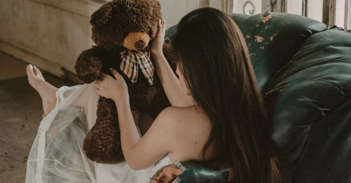 Una mujer se sienta en un sillón desgastado, sosteniendo un oso de peluche en un ambiente nostálgico en interiores.