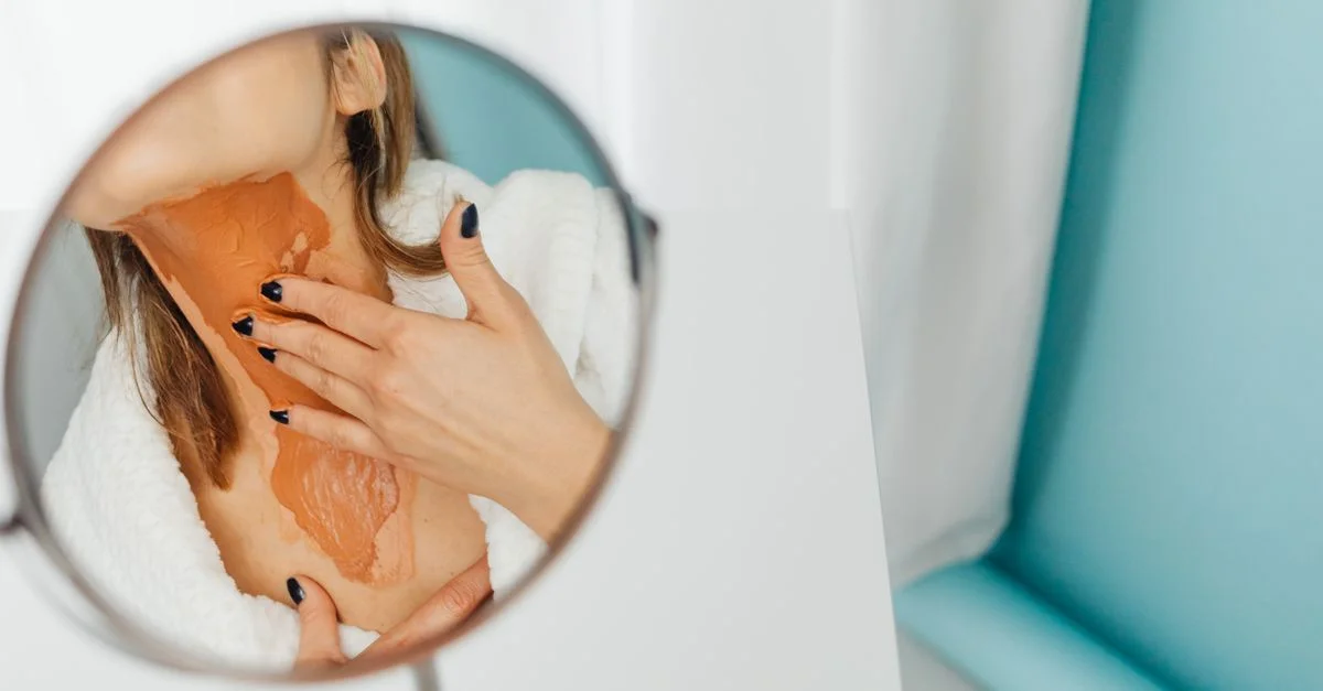 Primer plano de una mujer aplicando una capa gruesa de base en su cuello en un reflejo en el espejo.
