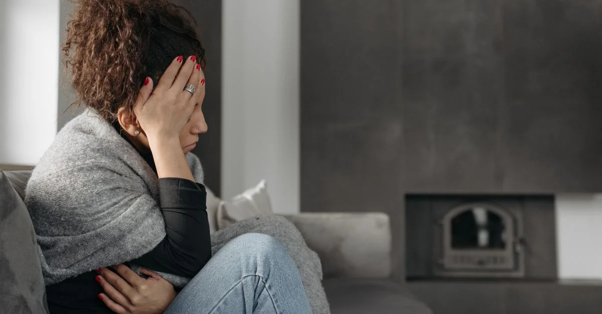 Una mujer está sentada en un sofá dentro de casa, envuelta en una manta, sosteniendo su cabeza con incomodidad.