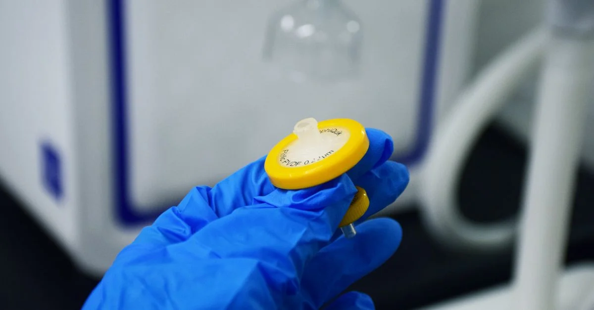 Primer plano detallado de una mano con guante azul sosteniendo un filtro de jeringa de laboratorio en un ambiente interior.