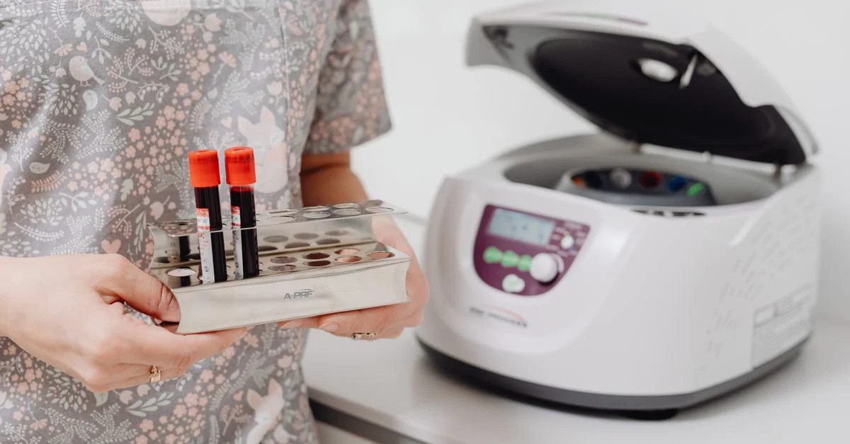 Profesional de salud sosteniendo frascos de sangre junto a una centrífuga de laboratorio, en interiores.